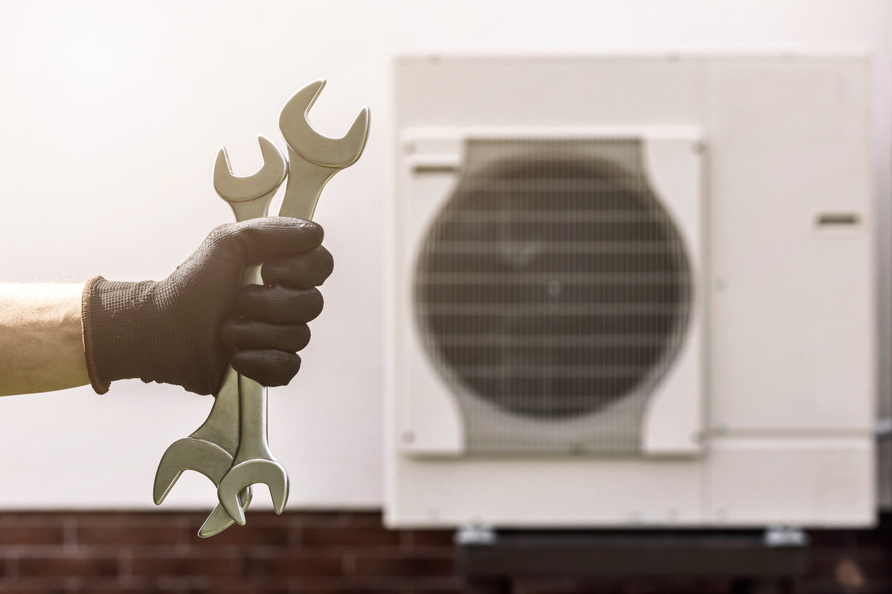 hand holding two spanners in front of heat pump to represent heat pump installation and maintenance